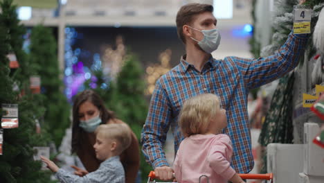 A-father-and-mother-with-two-children-son-and-daughter-are-choosing-Christmas-Tree-toys-and-home-decorations-on-Christmas-Eve-during-the-pandemic-and-quarantine.-family-in-medical-masks-in-the-store
